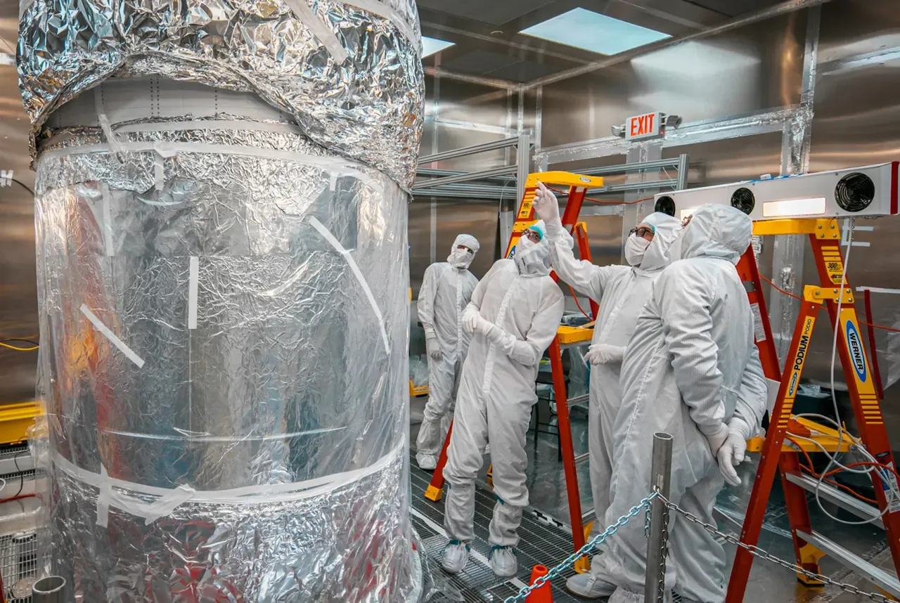 The LUX-ZEPLIN central detector wrapped in foil in a surface cleanroom, with four scientists in cleanroom suits inspecting it