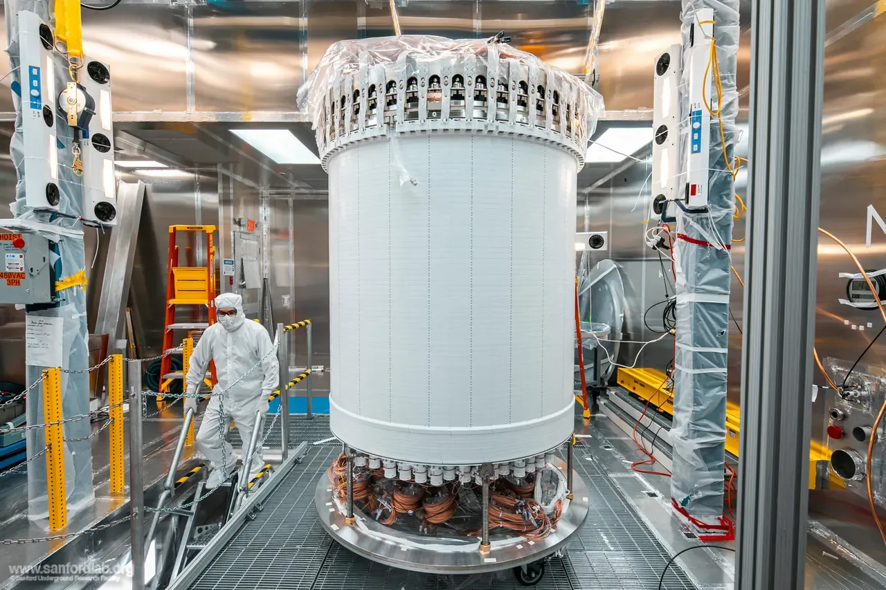 The white cylindrical LZ time projection chamber standing on a platform in a cleanroom, with a scientist in a cleanroom suit on the steps beside it